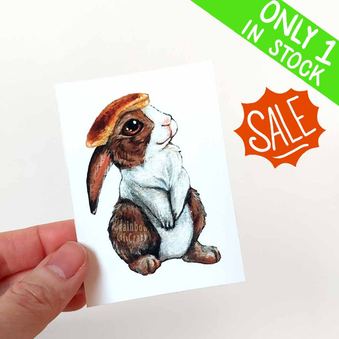 an aceo of a brown and white Dutch rabbit with a pancake on its head 