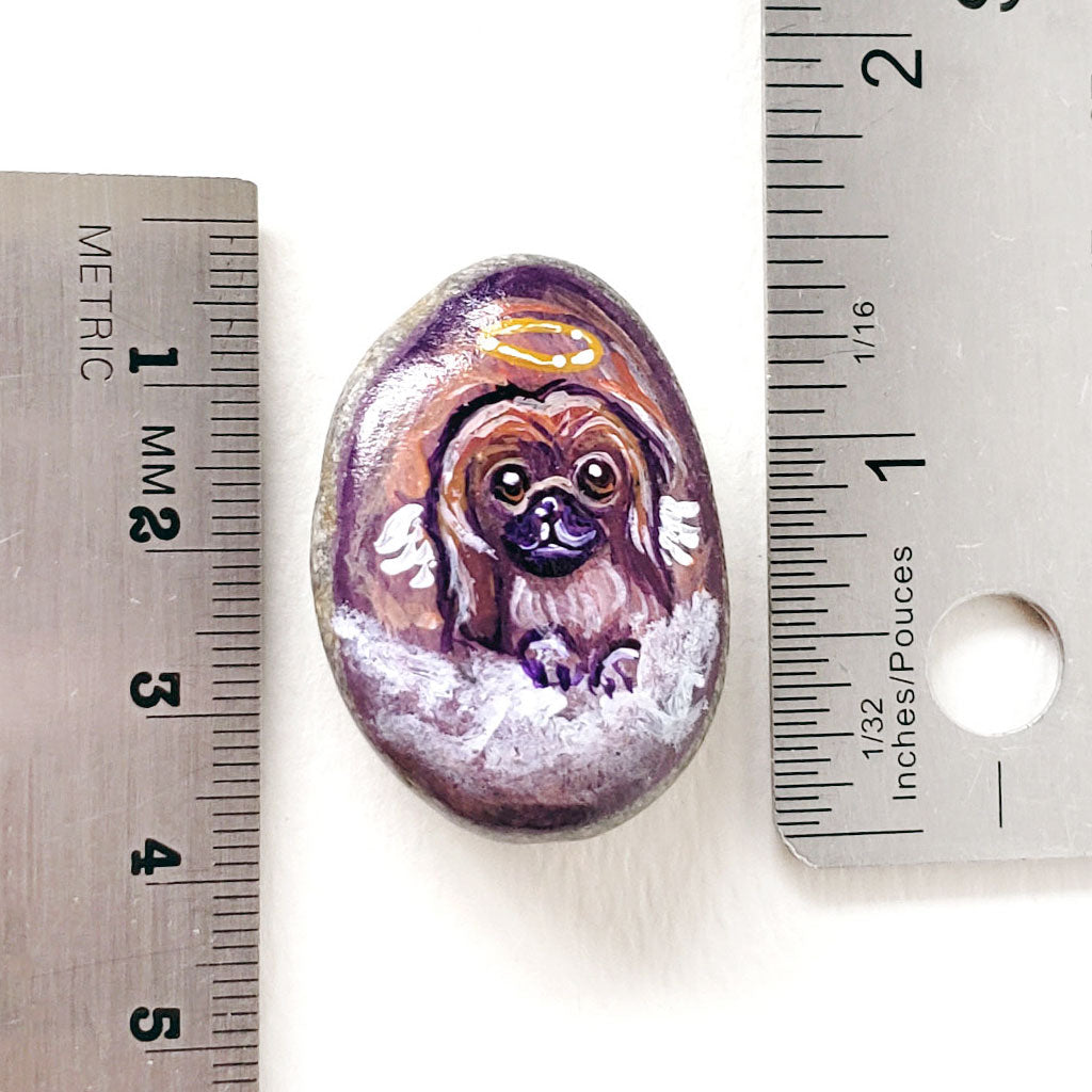 A small beach stone painted with a portrait of a Pekingese dog as an angel, next to two rulers to show its size: 1 7/16