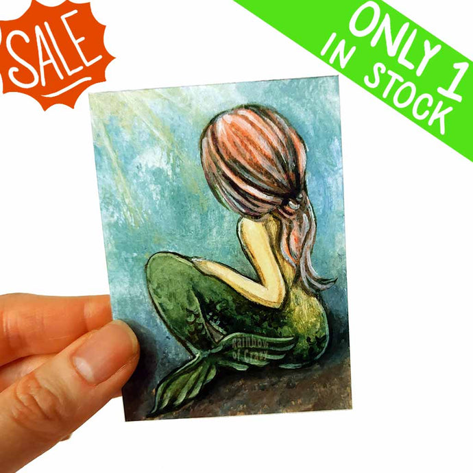 an aceo featuring a mermaid, face hidden with long pink hair, sitting on the bottom of the ocean floor.  