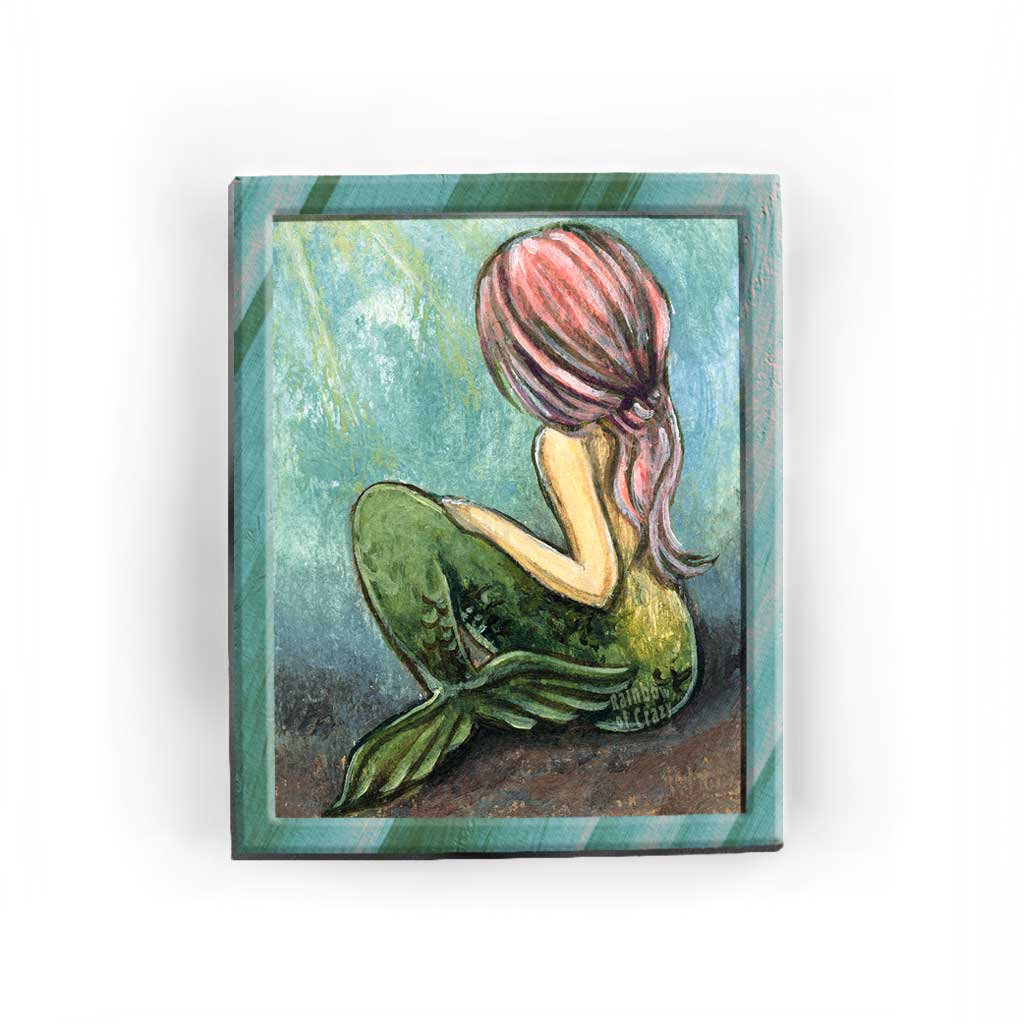 An art print of a mermaid sitting on the ocean floor, looking away from the viewere, as long pink hair flows down her back