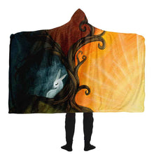 Load image into Gallery viewer, a man wears the Rabbit Tree of Life Hooded Blanket. It features an illustration of a white rabbit resting under the left side of a curly tree. A dark, stormy sky surrounds the sad bunny. But the right side of the tree reveals a beautiful sunrise. 
