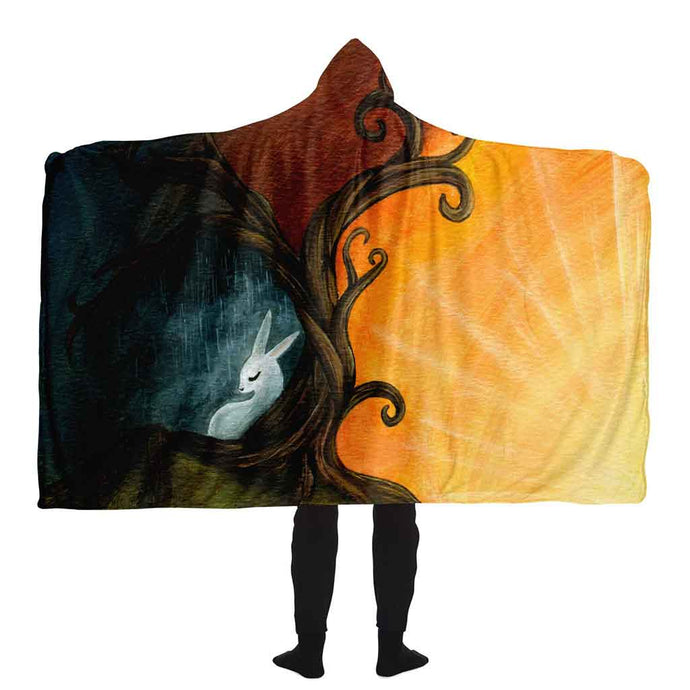 a man wears the Rabbit Tree of Life Hooded Blanket. It features an illustration of a white rabbit resting under the left side of a curly tree. A dark, stormy sky surrounds the sad bunny. But the right side of the tree reveals a beautiful sunrise. 