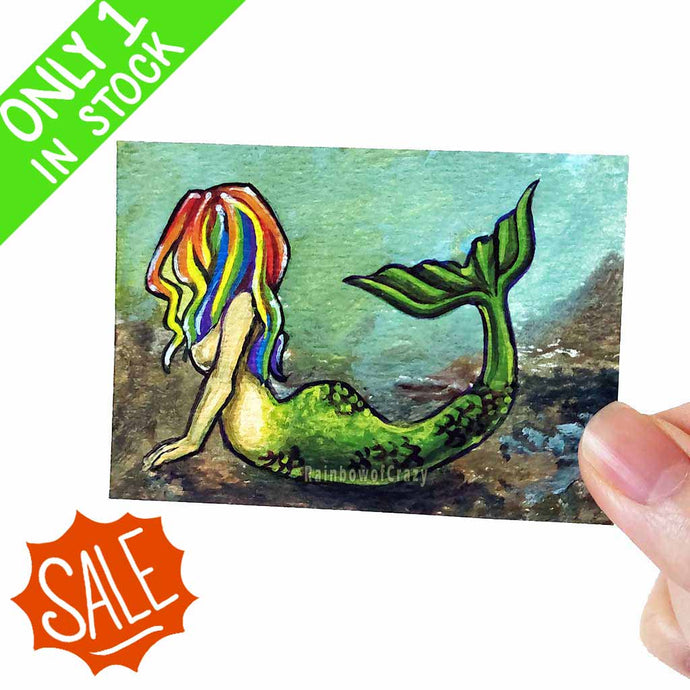 an aceo art print features art of a little mermaid with beautifully bright rainbow hair. She lies on the ocean floor, arching her back and raising her tail