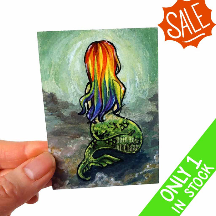 an aceo featuring art of a mermaid sitting on the ocean floor, with rainbow hair, facing away from the viewer