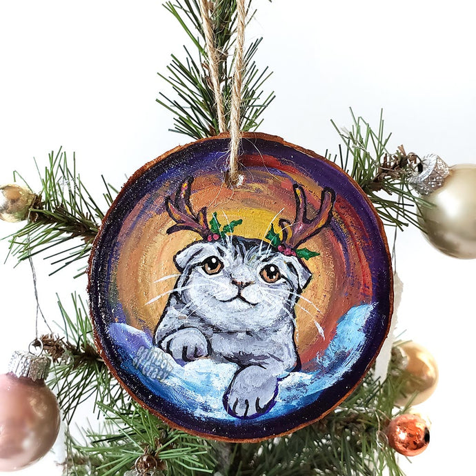 A wood Christmas ornament, hand painted with a portrait of a Scottish fold cat with reindeer antlers, sitting on the snow.