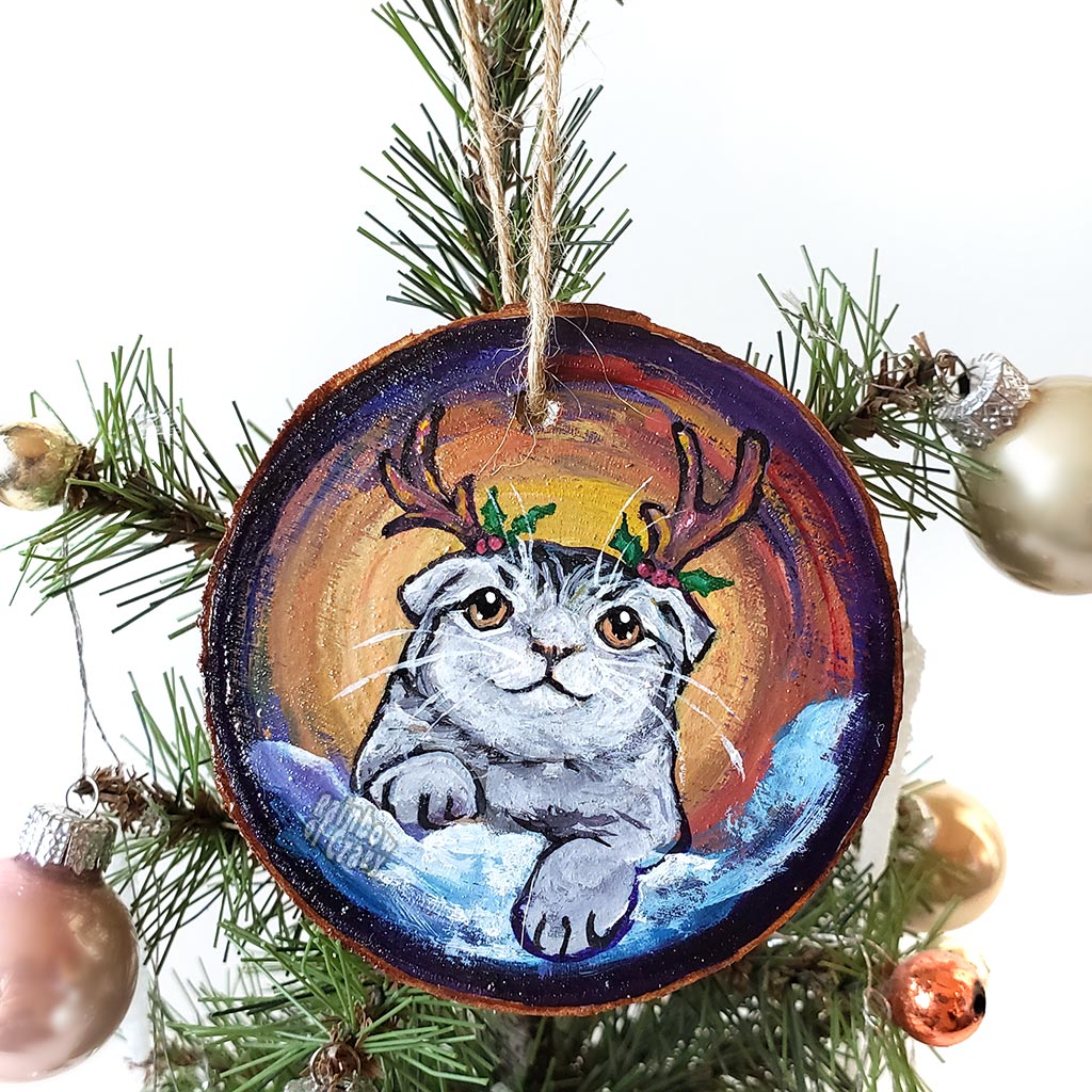 A wood Christmas ornament, hand painted with a portrait of a Scottish fold cat with reindeer antlers, sitting on the snow.