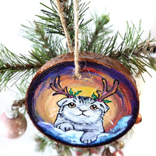 Load image into Gallery viewer, A wood Christmas ornament, hand painted with a portrait of a Scottish fold cat with reindeer antlers, sitting on the snow.
