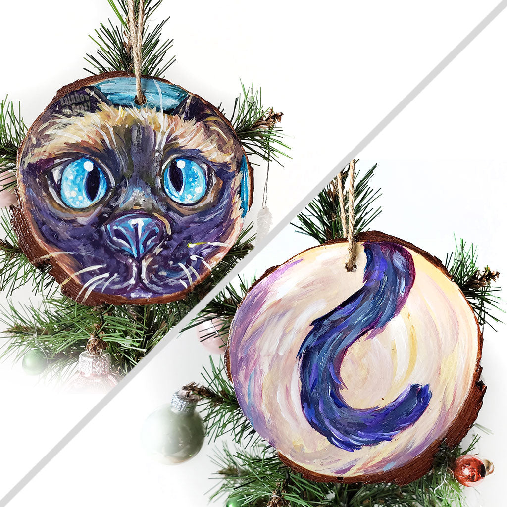 A wood ornament featuring art of a siamese cat's face with blue eyes, and it's tail on the back.