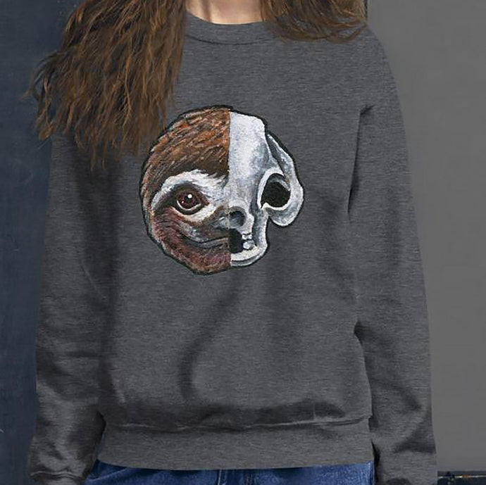 A woman wears a unisex sweatshirt in the colour dark heather grey, printed with a graphic of a split image: the left side features a sloth's face, and the right side features an evil looking sloth skull.