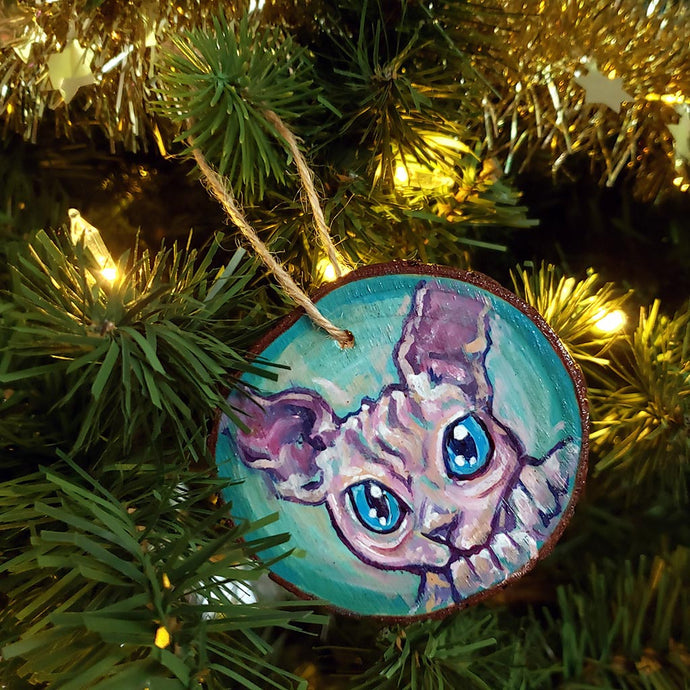 A handmade Christmas tree wood ornament, featuring a painting of a sphynx cat with blue eyes