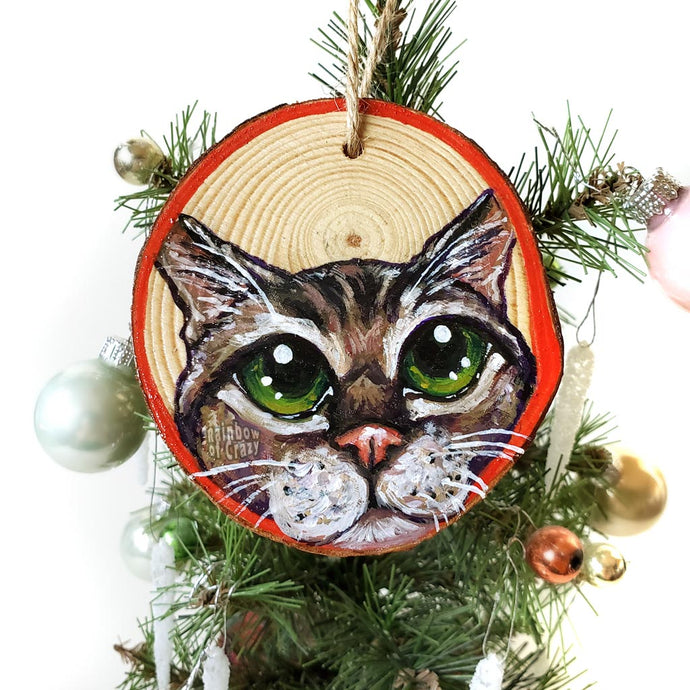 A Christmas wood ornament, hand painted with the face of a tabby cat with green eyes, and a red border.
