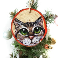 A Christmas wood ornament, hand painted with the face of a tabby cat with green eyes, and a red border.