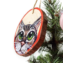 Load image into Gallery viewer, A Christmas wood ornament, features a painting of the face of a tabby cat with green eyes, and a red border.
