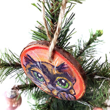 Load image into Gallery viewer, A wood Christmas tree ornament, hand painted with art of a tortoiseshell cat, with green eyes, in front of a red and pink background
