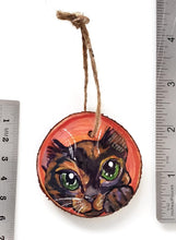 Load image into Gallery viewer, A wood Christmas tree ornament, hand painted with art of a tortoiseshell cat, with green eyes, in front of a red and pink background
