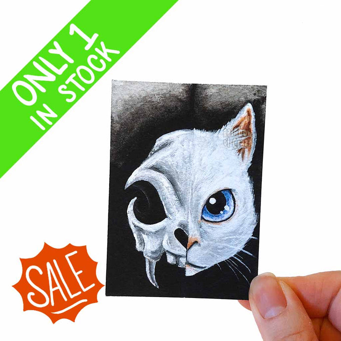 This macabre ACEO art print features a white cat with blue eyes of the left side, while the other has a scary cat skull. 