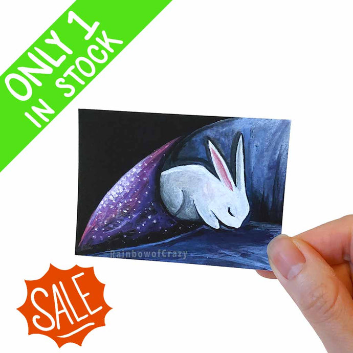 This mini art print features a white rabbit sleeping, with the corner of the image wrapped around itself like a blanket covered in stars. 