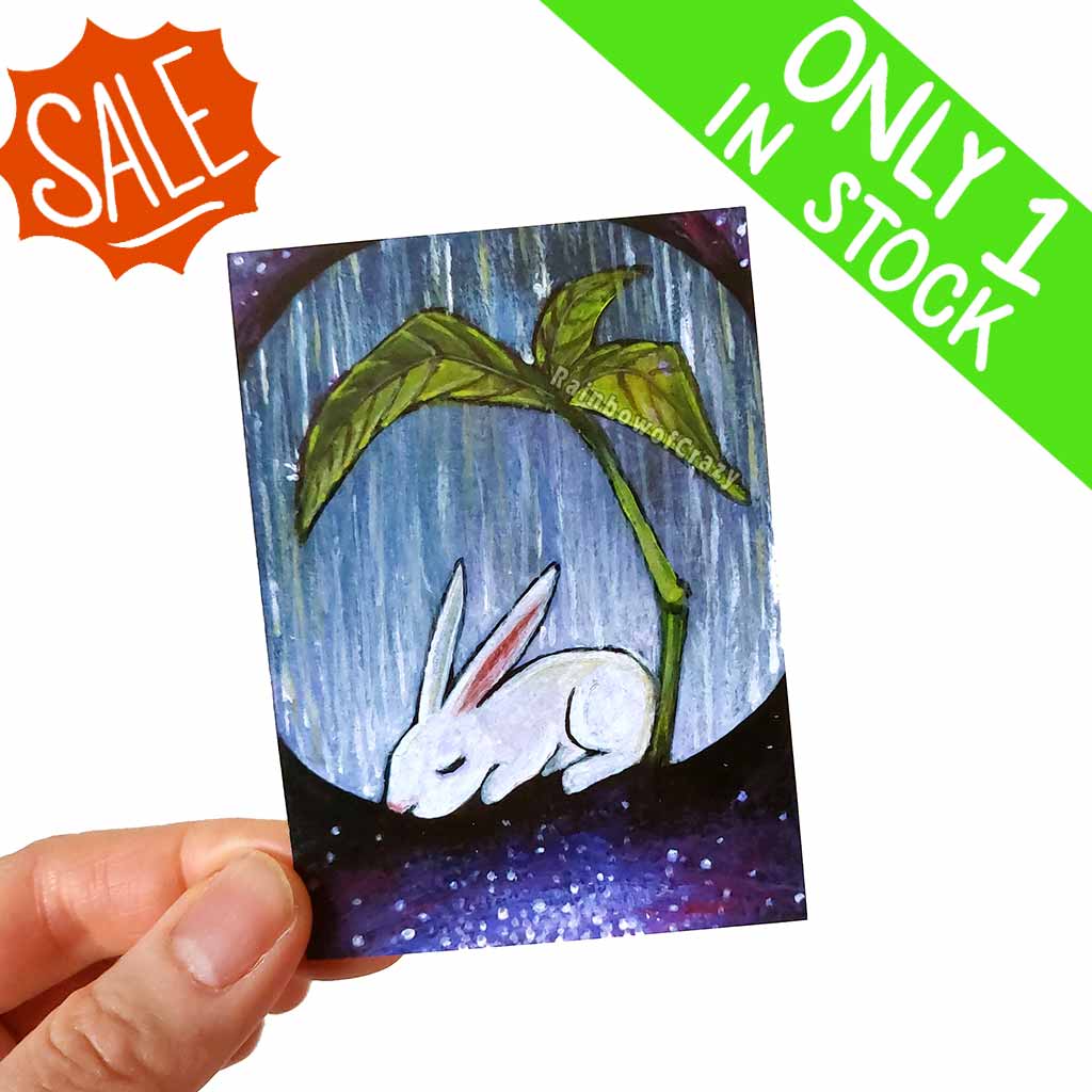 an aceo features a white rabbit resting under a giant leaf, as heavy rain falls. The ground fades into a galaxy of stars. 