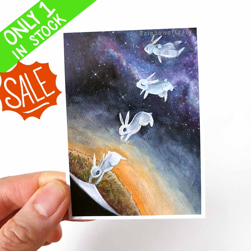 ACEO art print features a white rabbit holding onto a corner of the earth before letting go and floating into a galaxy of stars. 