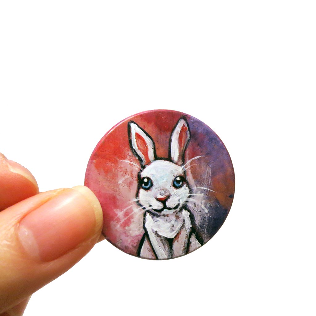A circle wood disc painted with art of a white rabbit with blue eyes, available as a keepsake or necklace
